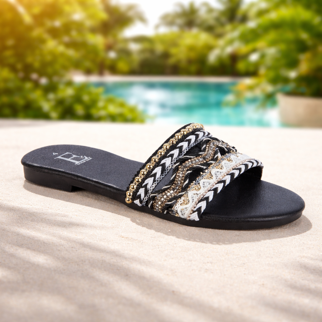 Black sandal with decorative straps on a sandy surface with a pool and greenery 